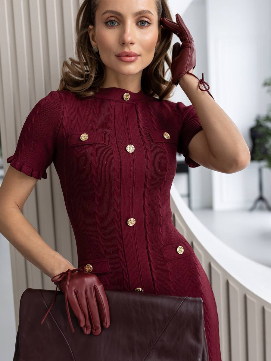 Woman wearing a burgundy dress with gold buttons, holding a matching handbag indoors.