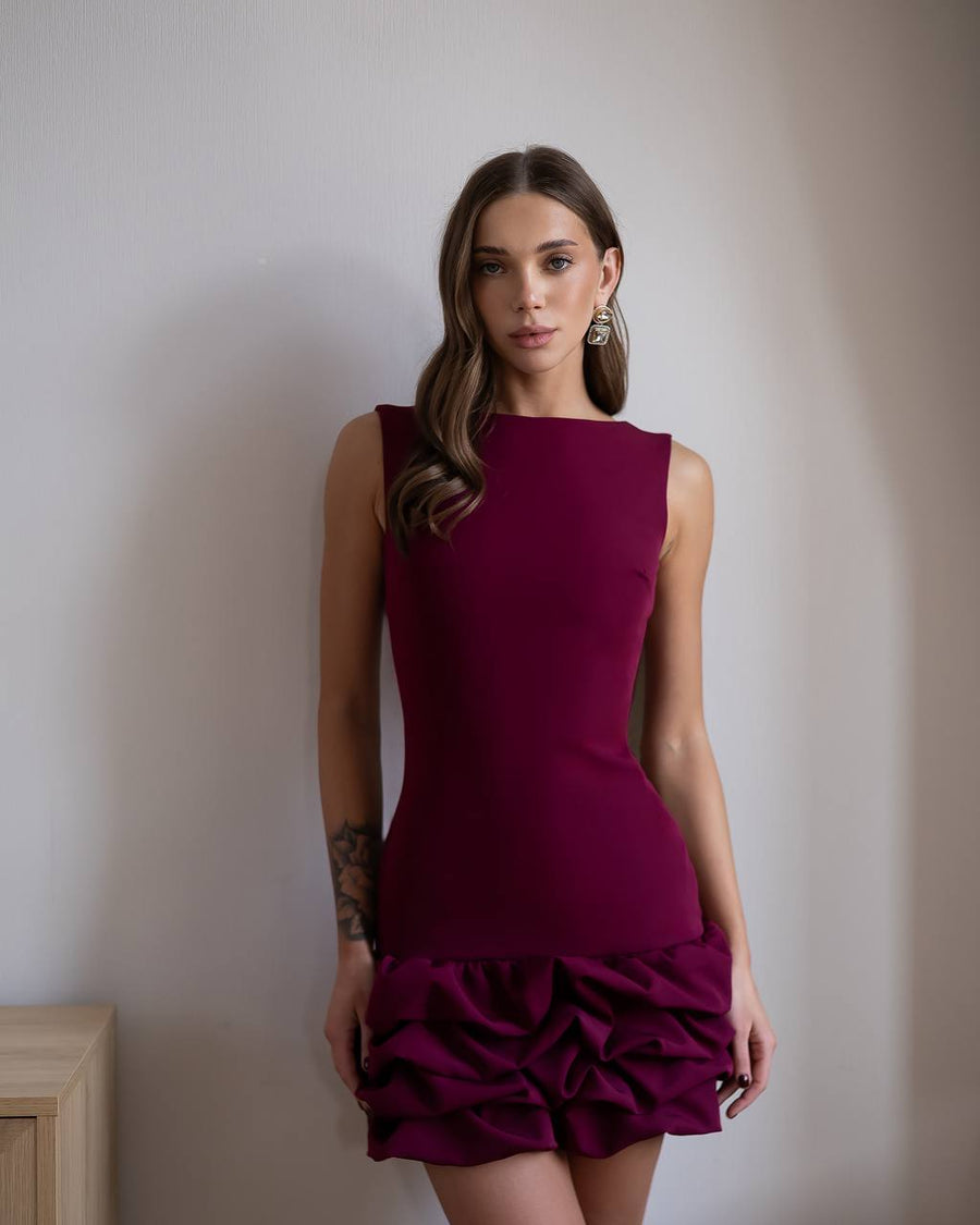 Woman wearing a burgundy dress with a ruffled skirt against a plain wall.