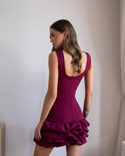 Woman wearing a burgundy dress with a ruffled skirt against a plain wall.