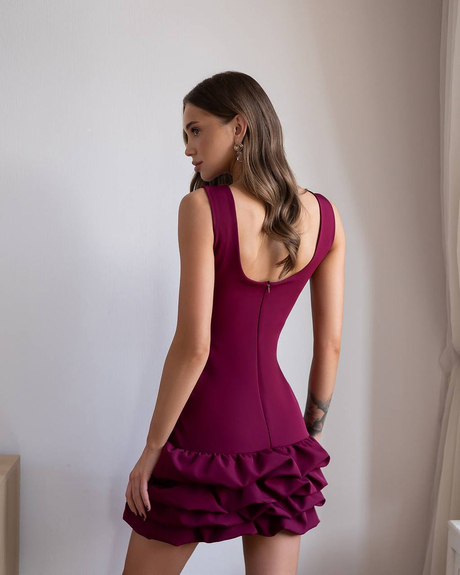 Woman wearing a burgundy dress with a ruffled skirt against a plain wall.