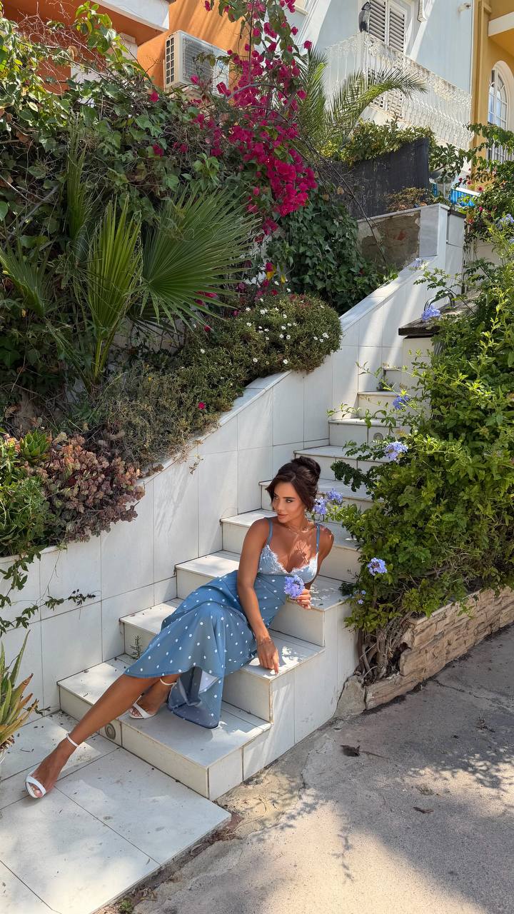 Woman in a blue dress sitting on stone steps surrounded by greenery and flowers.