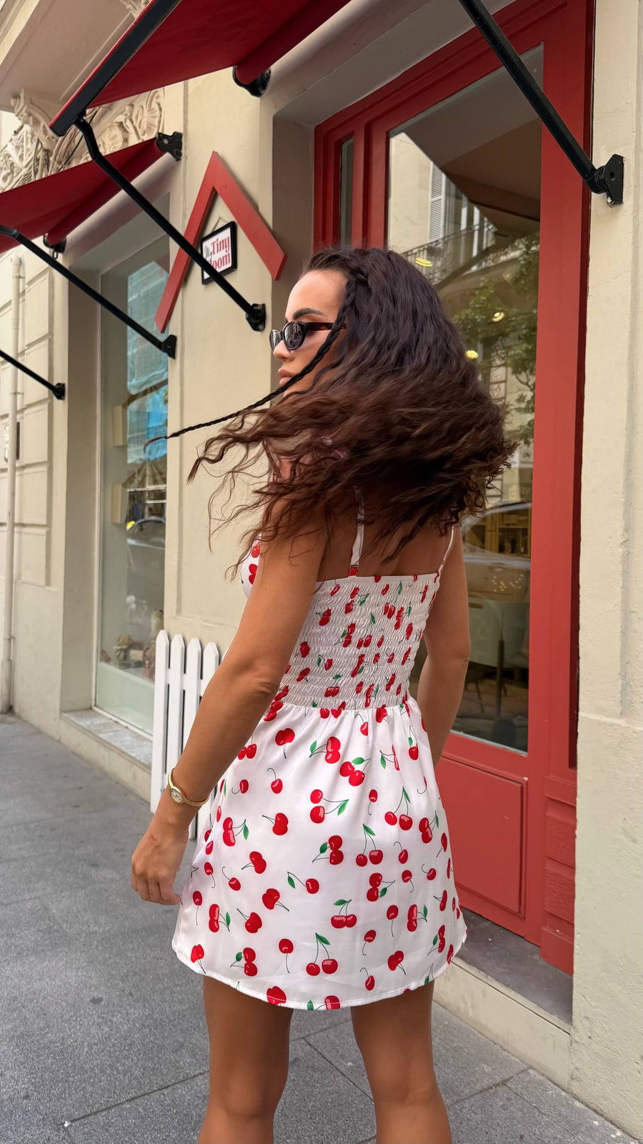 Woman in a white dress with red cherries standing on a sidewalk in front of a store with a red door.