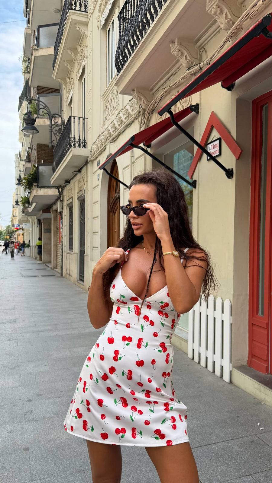 A woman wearing a white mini dress with a red cherry print stands on a city sidewalk, adjusting black sunglasses. The background shows classic European-style buildings with balconies and awnings.