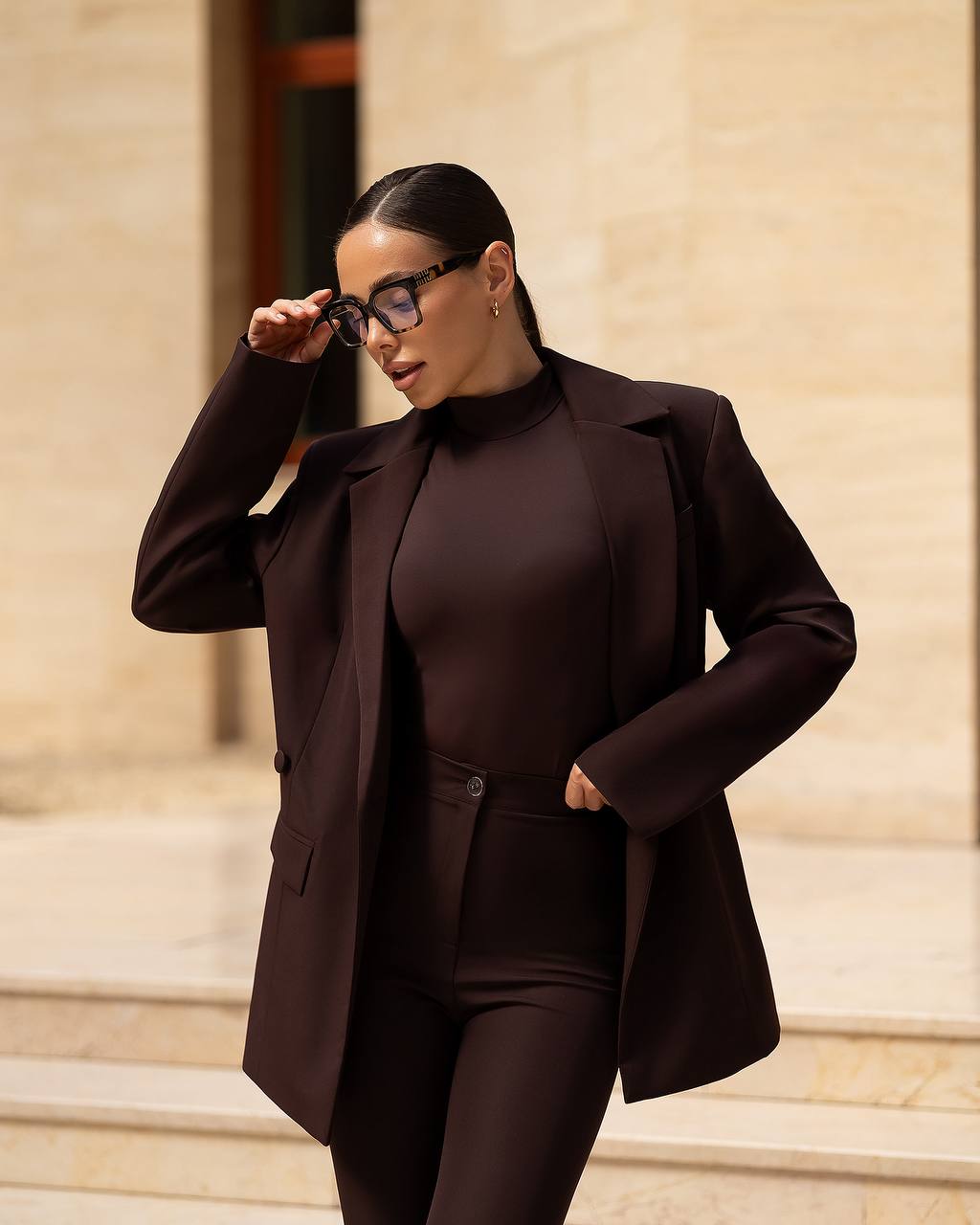 Woman in a dark brown suit standing on steps with a beige wall background