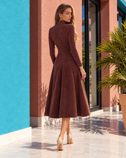 A woman stands outdoors on a tiled walkway wearing a long-sleeve, high-neck, knee-length brown dress with a flared skirt, paired with beige high heels. The background features a bright blue wall, a terracotta-colored building column, and a potted palm.