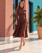 A woman models a long-sleeve, high-neck, knee-length brown dress with a flared skirt, paired with beige pointed-toe heels, set against a modern architectural background with pink and teal walls.