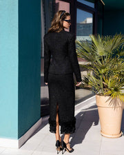 A woman stands outdoors wearing a black textured long-sleeve jacket and matching black skirt with ruffled hems, paired with black heels. She is photographed from the back beside a teal wall, large windows, and a potted palm.