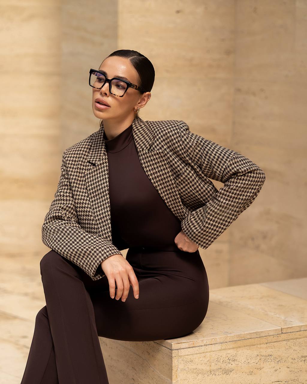 Woman wearing a checkered blazer over a brown top and pants, sitting on a stone bench.