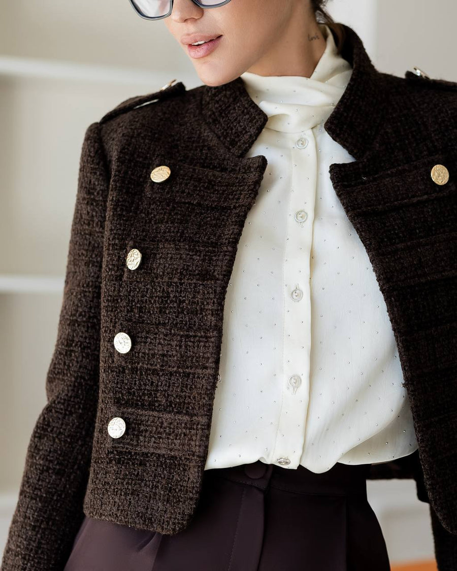 A close-up of a person wearing a dark brown textured tweed jacket with gold buttons over a white polka-dot blouse and dark trousers.