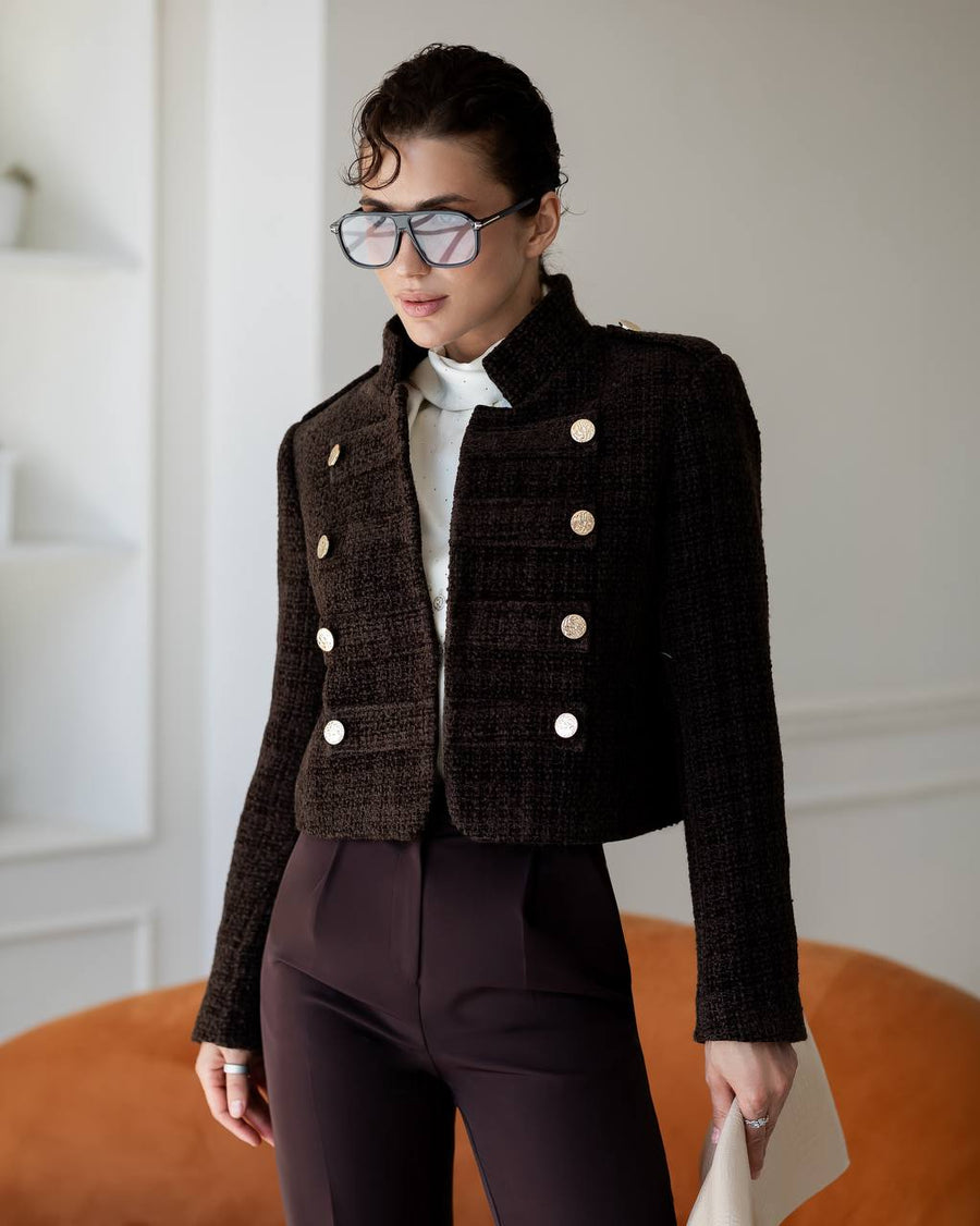 A woman wearing a dark brown tweed double-breasted jacket with gold buttons, a white shirt, and dark brown trousers, standing in a bright indoor setting.