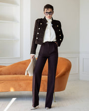 A model stands in a bright room wearing a dark brown tweed double-breasted jacket, a white ruffled blouse, dark plum trousers, and holding a beige clutch. An orange velvet sofa is in the background.