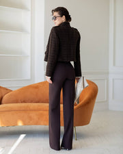 A woman stands with her back to the camera in a bright room, wearing a dark brown tweed cropped jacket and dark plum wide-leg trousers, standing in front of an orange velvet sofa.