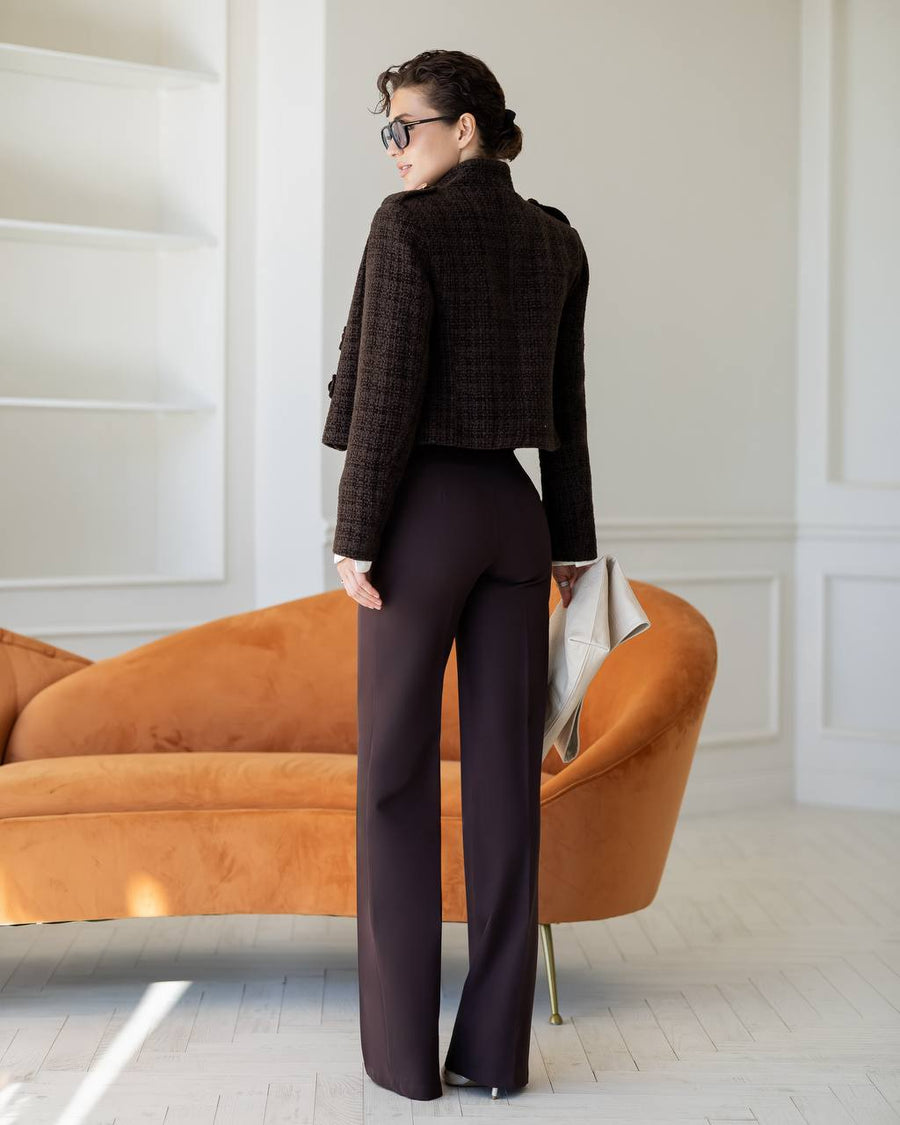 A woman stands with her back to the camera in a bright room, wearing a dark brown tweed cropped jacket and dark plum wide-leg trousers, standing in front of an orange velvet sofa.
