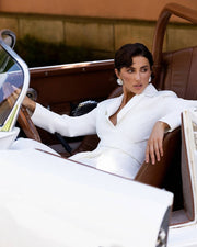 A woman wearing a white tailored blazer and matching trousers sits in a brown leather convertible car, with the car's white exterior visible in the foreground.