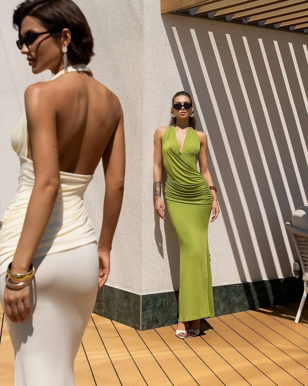 Two women in elegant dresses standing on a wooden deck with a striped wall in the background.