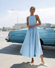 Woman in a light blue dress standing next to a vintage blue car on a sunny day.
