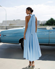 Woman in a light blue dress standing next to a vintage car on a sunny day.