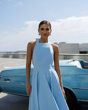 Woman in a light blue dress standing next to a vintage car with a clear sky background