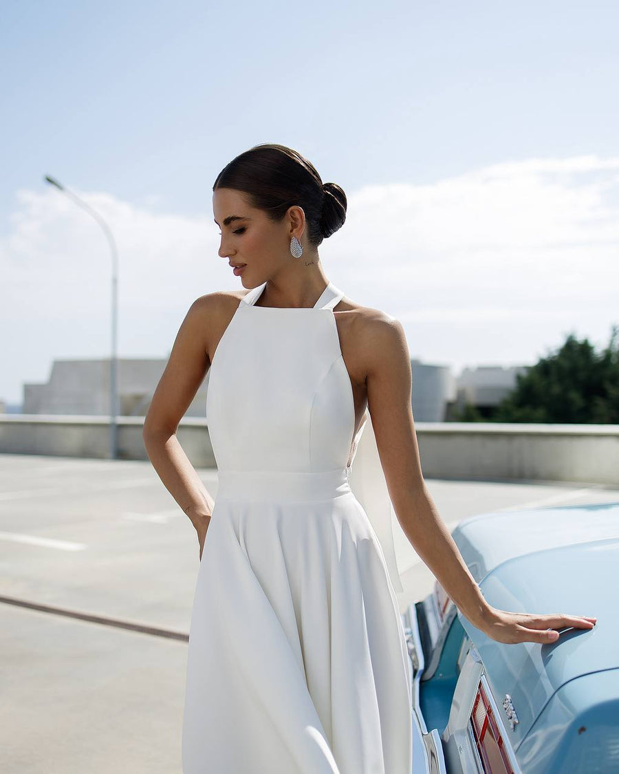 Woman in a white dress standing next to a vintage car on a sunny day.