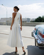 Woman in a white dress standing next to a vintage car on a road.