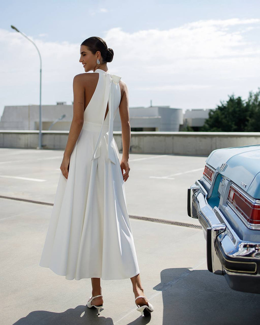 Woman in a white dress standing next to a vintage car on a road.