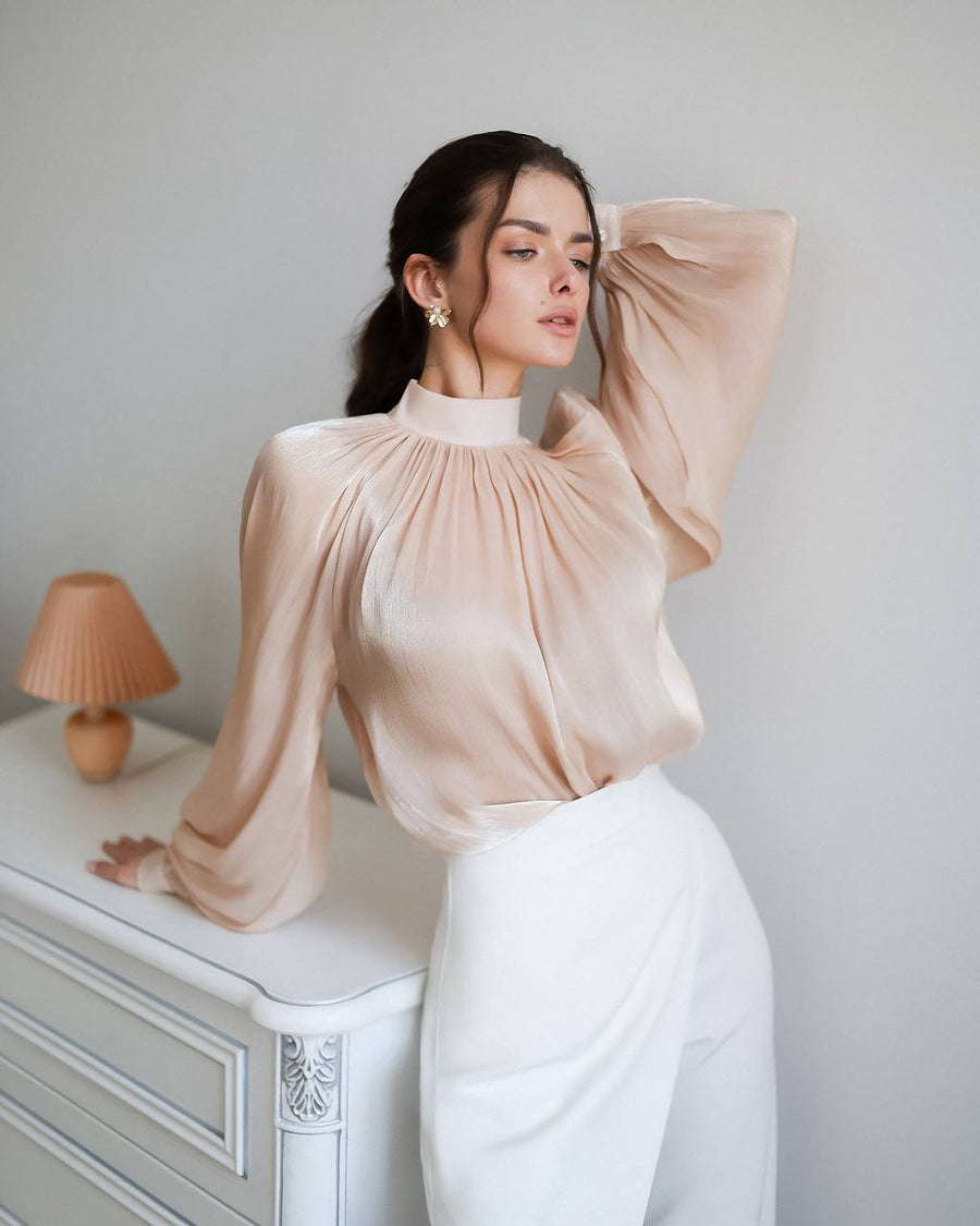 Woman wearing a beige blouse and white pants in a minimalistic room.