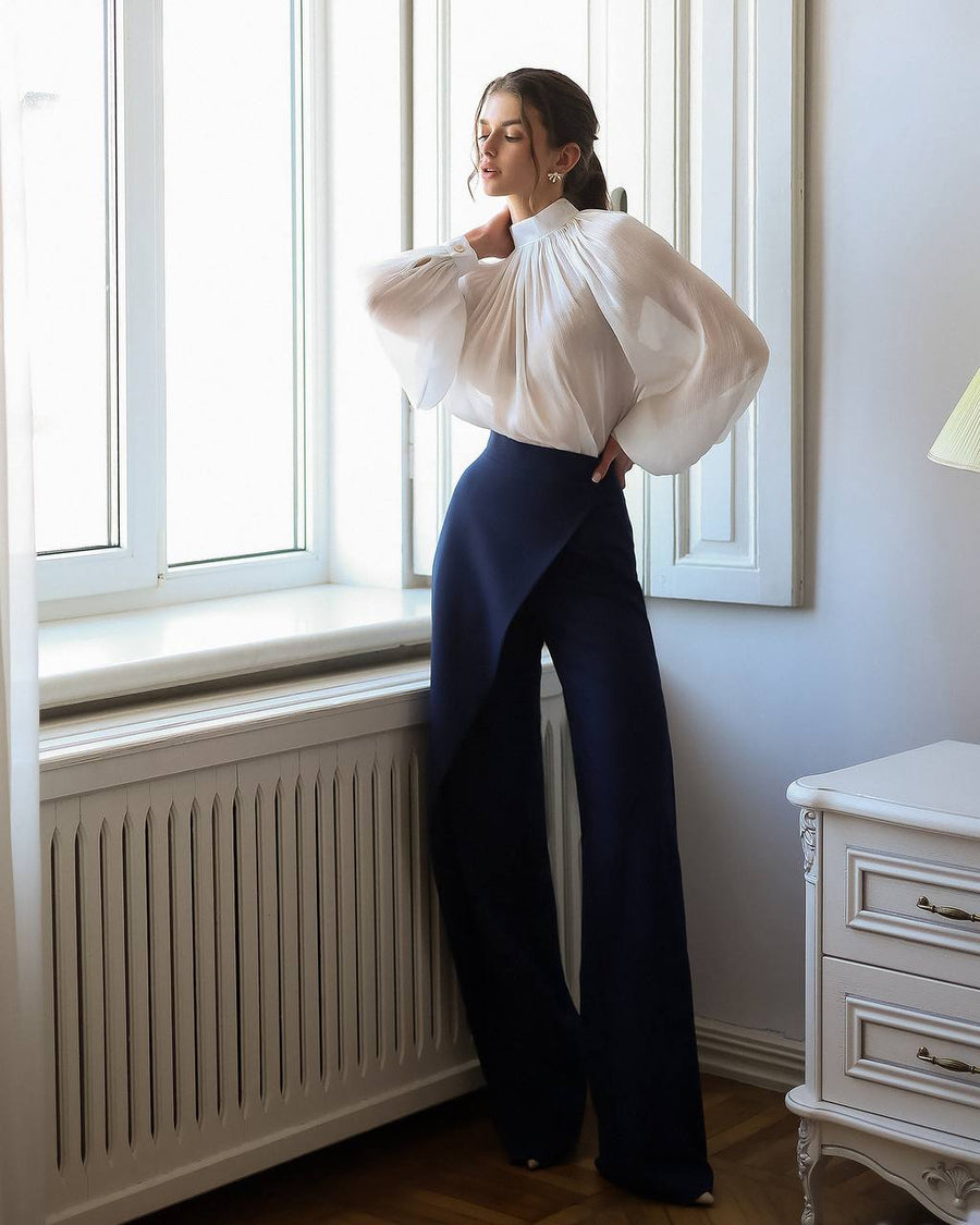 Woman wearing a white blouse and dark blue pants standing by a window in a room with a radiator and dresser.