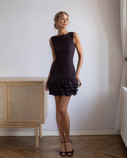 Woman wearing a brown dress standing in a room with wooden flooring and a light-colored wall.