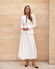 Woman wearing a white suit standing against a beige wall.