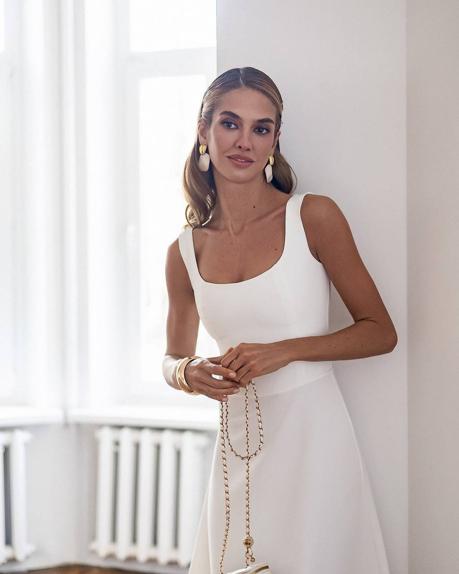 Woman in a white dress standing in a bright room with large windows.
