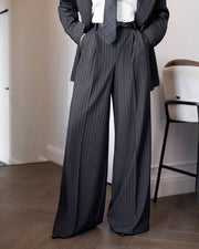 A person wearing dark gray pinstripe wide-leg trousers with a matching blazer, white shirt, and tie, standing in an indoor room with wood flooring and a chair.