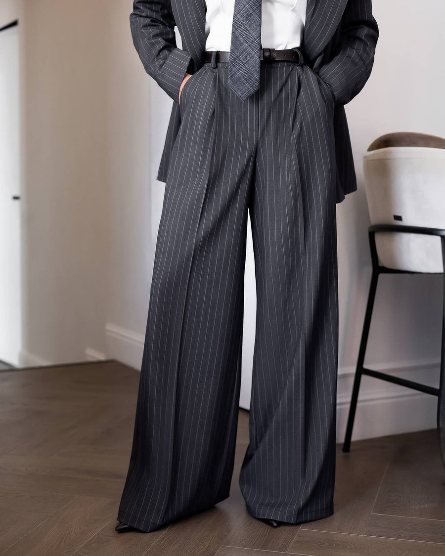 A person wearing dark gray pinstripe wide-leg trousers with a matching blazer, white shirt, and tie, standing in an indoor room with wood flooring and a chair.