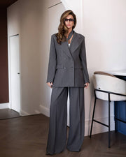 A woman modeling a dark gray double-breasted pinstripe suit with wide-leg trousers, styled with a white top and sunglasses in a modern indoor setting.