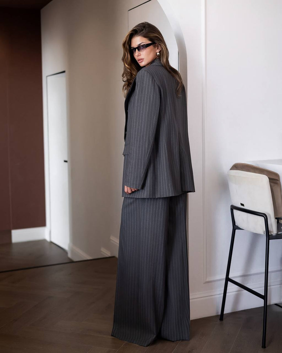 A woman wearing a dark gray pinstripe blazer and matching wide-leg trousers, styled with black sunglasses, standing in a minimalist interior setting.