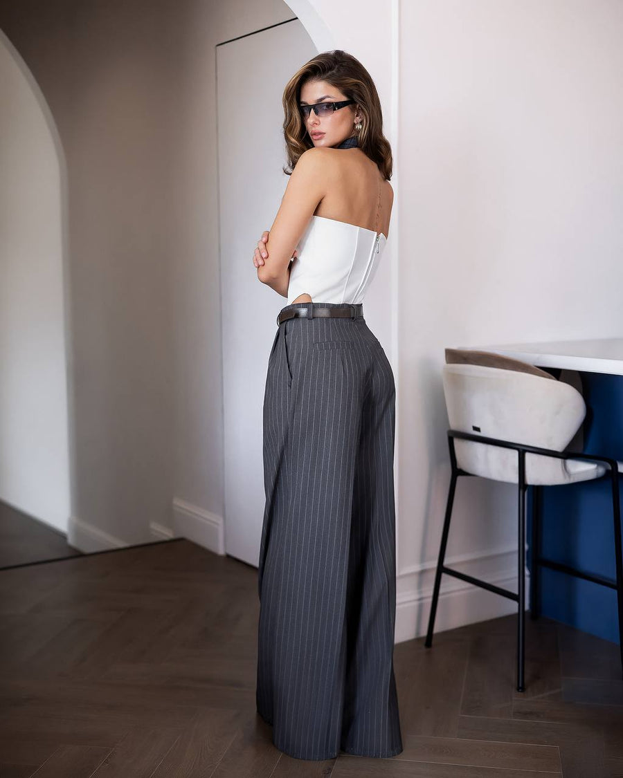 A woman stands in a doorway wearing a white strapless tube top and dark gray pinstriped wide-leg trousers with a black belt, photographed indoors with a chair and blue cabinet in the background.