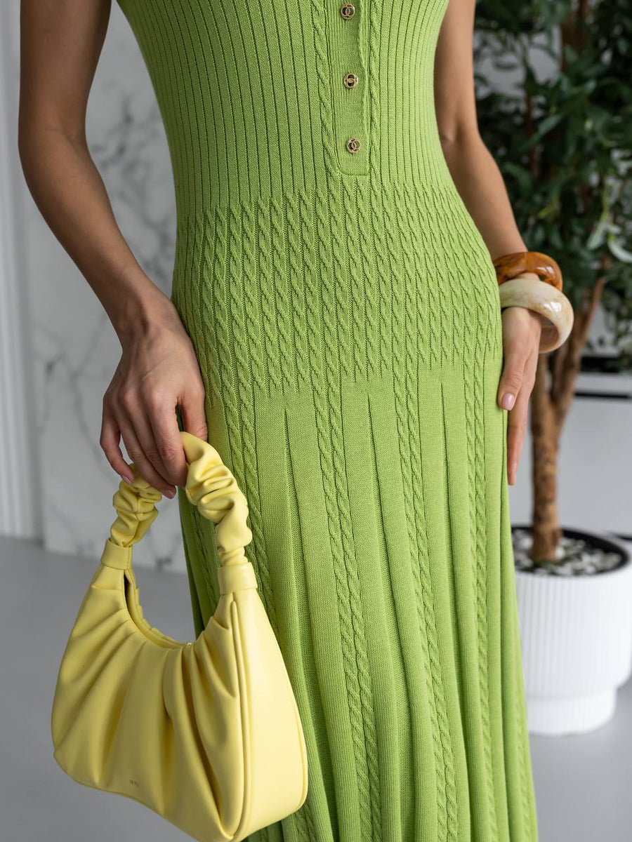 A woman wearing a lime green ribbed knit sleeveless dress with a buttoned neckline and holding a matching light yellow-green pleated shoulder bag. She is standing indoors near a potted plant.