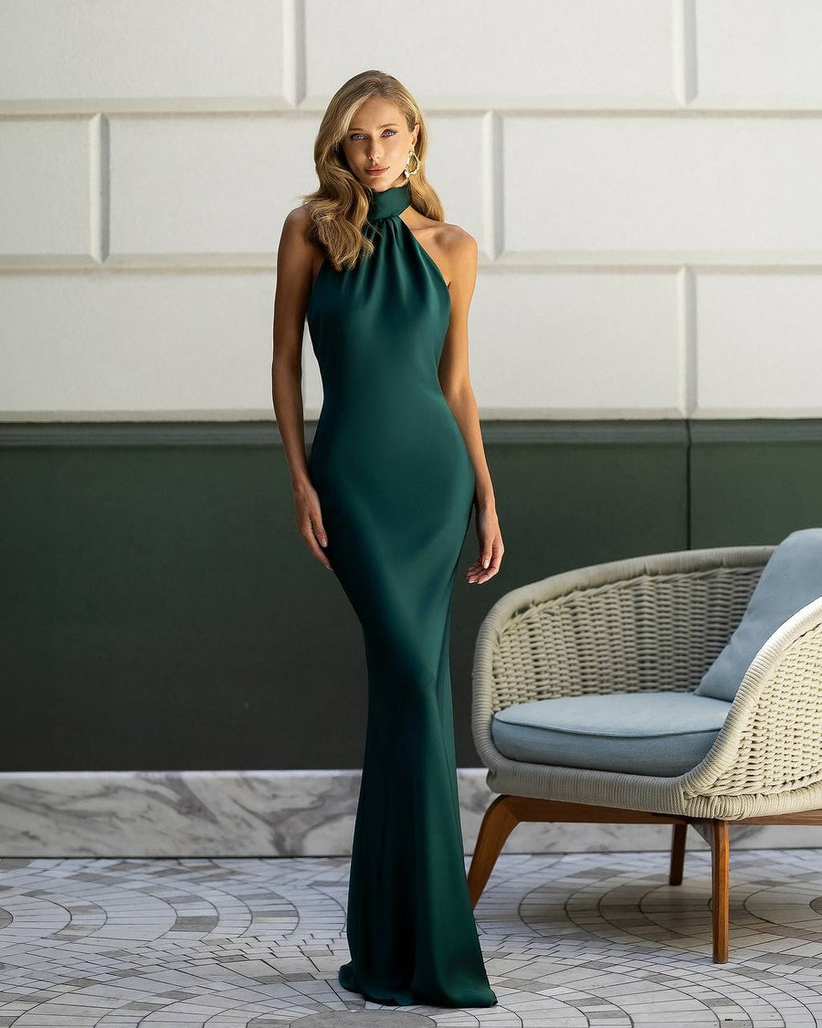 Woman in a green evening gown standing in a stylish room with a chair and decorative wall.