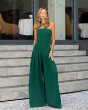 A woman models a strapless emerald green wide-leg jumpsuit, standing on stone steps in front of a modern building with large windows.