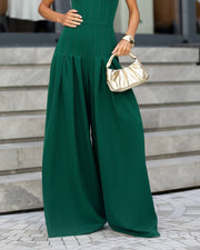 A woman wearing a dark emerald green wide-leg jumpsuit, holding a metallic gold handbag while standing on stone steps.
