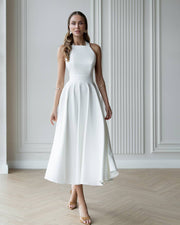 Woman wearing a white dress standing in a room with light-colored walls and wooden floor.