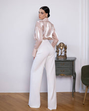 Woman wearing a sheer pink top and white pants standing in a room with a small table and chair.