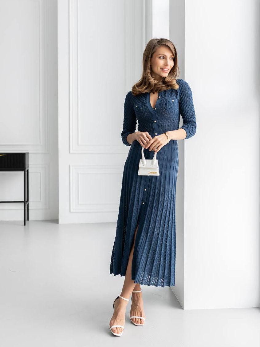 A woman wearing a navy blue textured knit midi dress with a button-up front and long sleeves, holding a small white handbag and wearing white heeled sandals, standing in a bright white room with classic paneled walls.