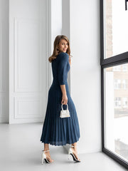 A woman wearing a navy blue textured midi dress with three-quarter sleeves and pleated detailing, holding a small white handbag and wearing white high-heeled sandals, standing in a bright room next to a large window.