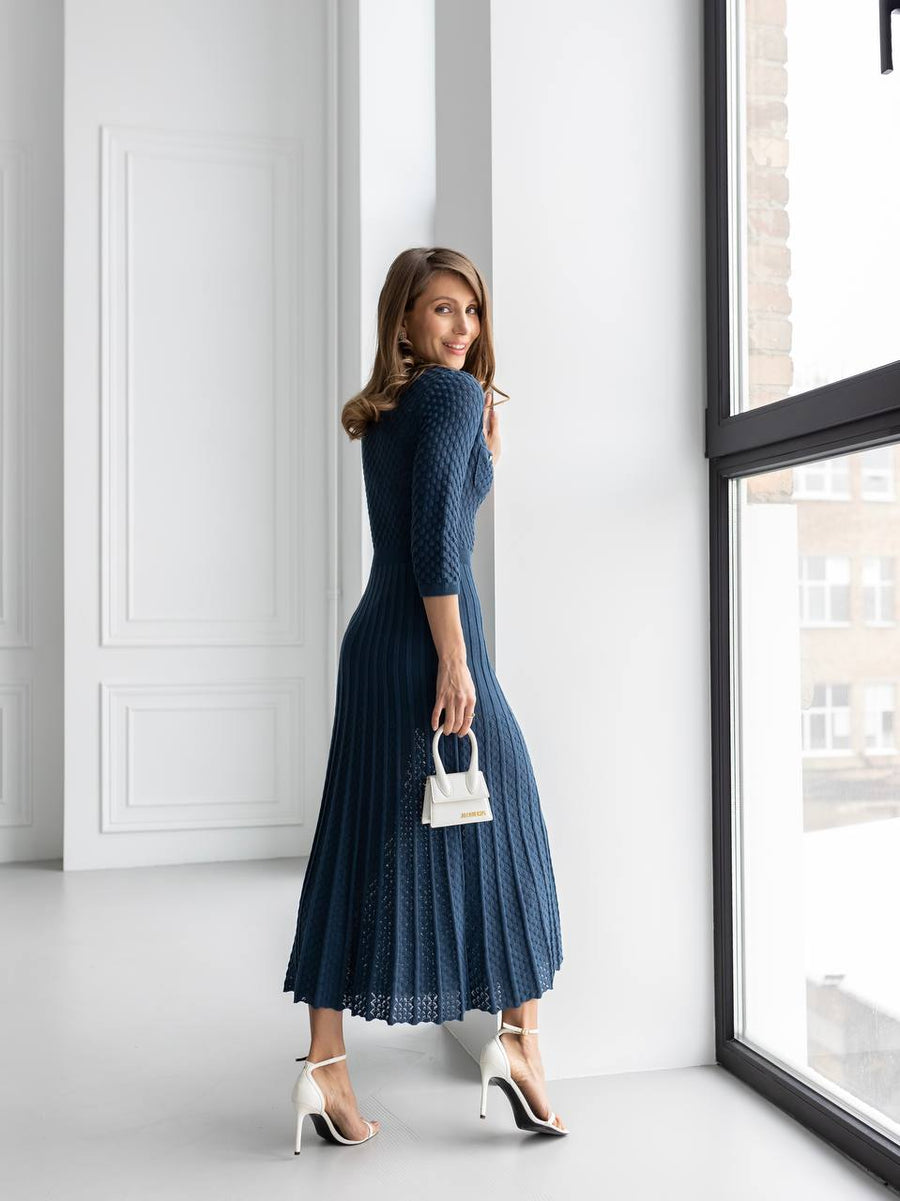 A woman wearing a navy blue textured midi dress with three-quarter sleeves and pleated detailing, holding a small white handbag and wearing white high-heeled sandals, standing in a bright room next to a large window.
