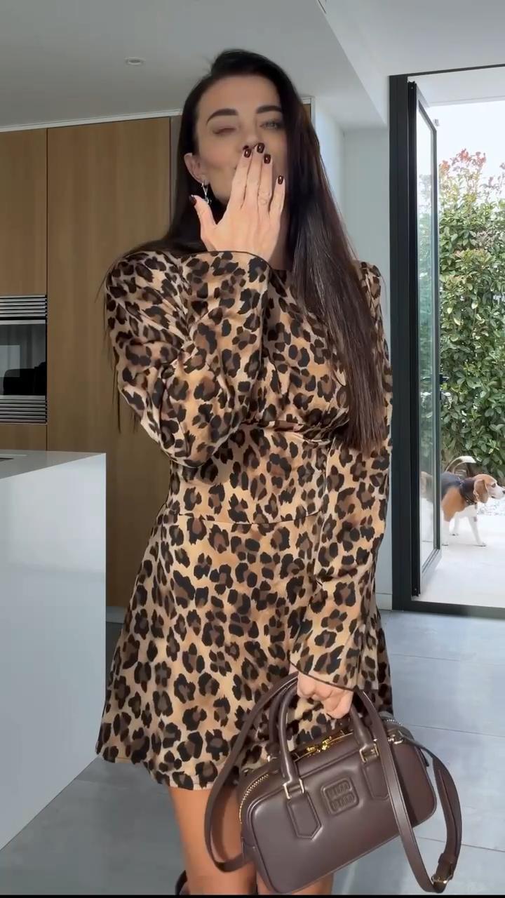 Woman in leopard print dress holding a brown handbag in a modern kitchen.