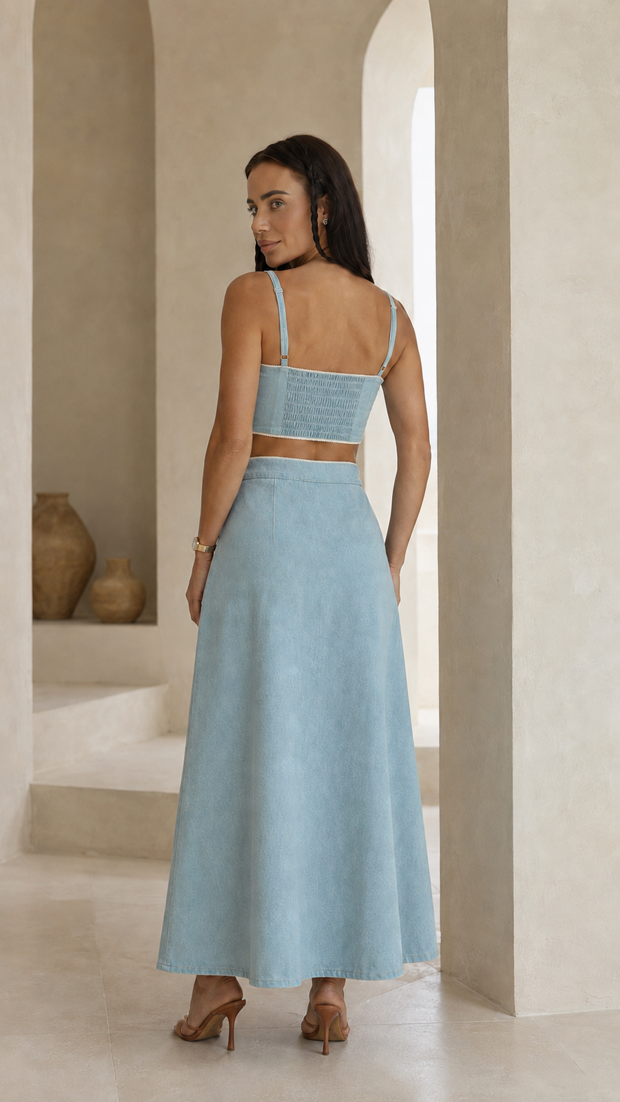 A woman stands in a softly lit architectural interior wearing a light blue two-piece outfit with a cropped camisole-style top and a long skirt, shown from the back.