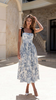 Woman in a blue floral dress standing outdoors near stone architecture.