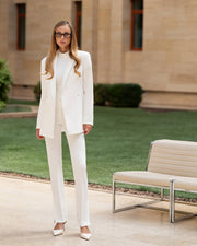 Woman in a white suit standing outdoors with a bench and building in the background