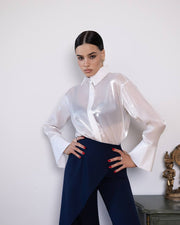 Woman wearing a sheer white blouse and navy pants against a white wall.