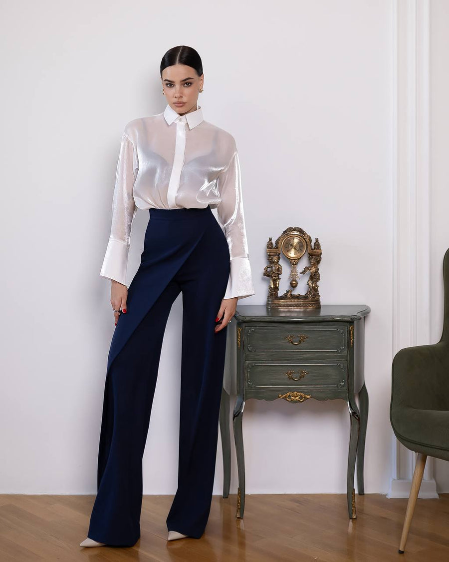 Woman wearing a white sheer blouse and navy pants standing in a room with a small table and chair.